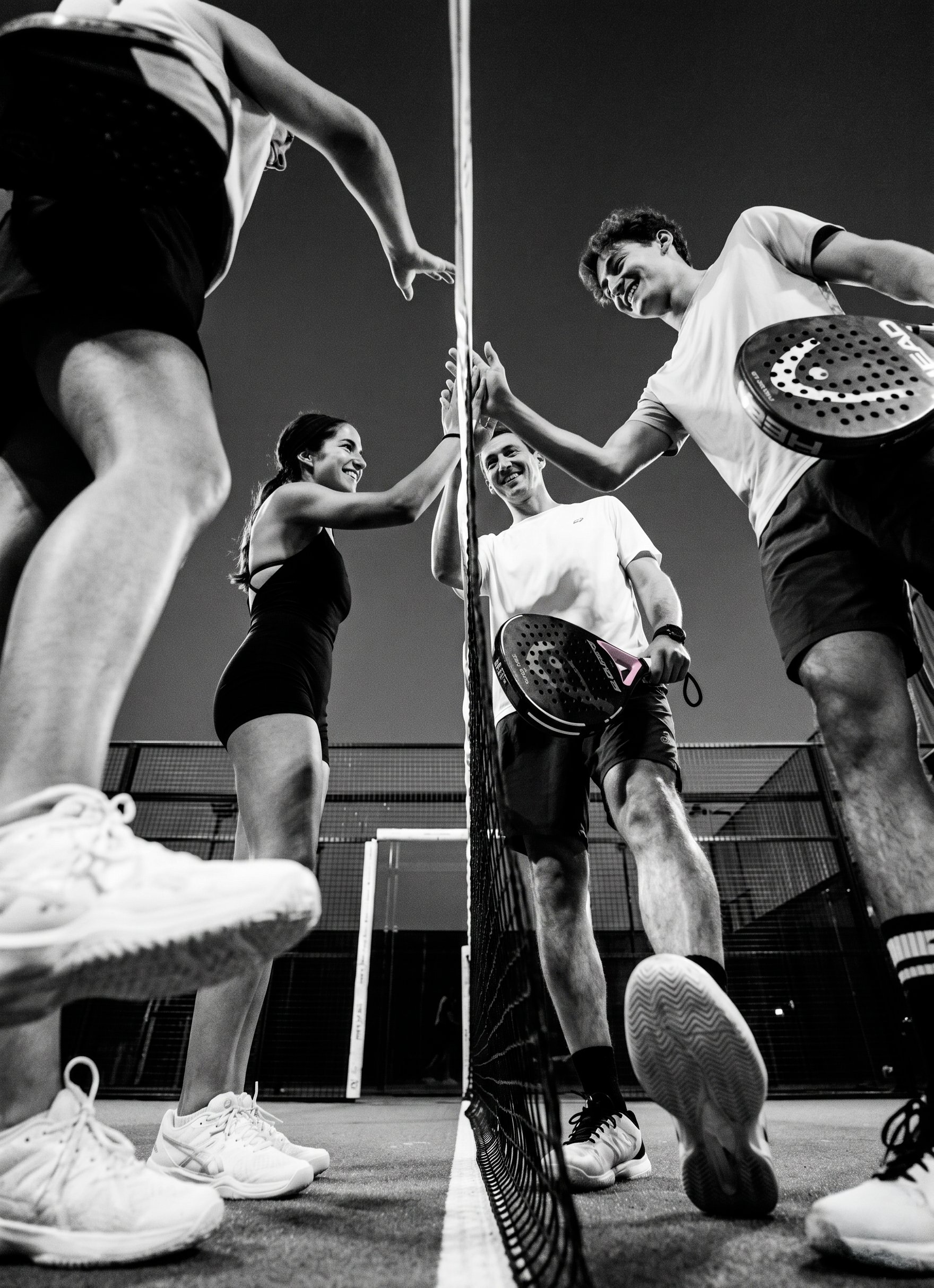 Four padel players celebrating across the net after a match — high-fives, smiles, paddles in hand.