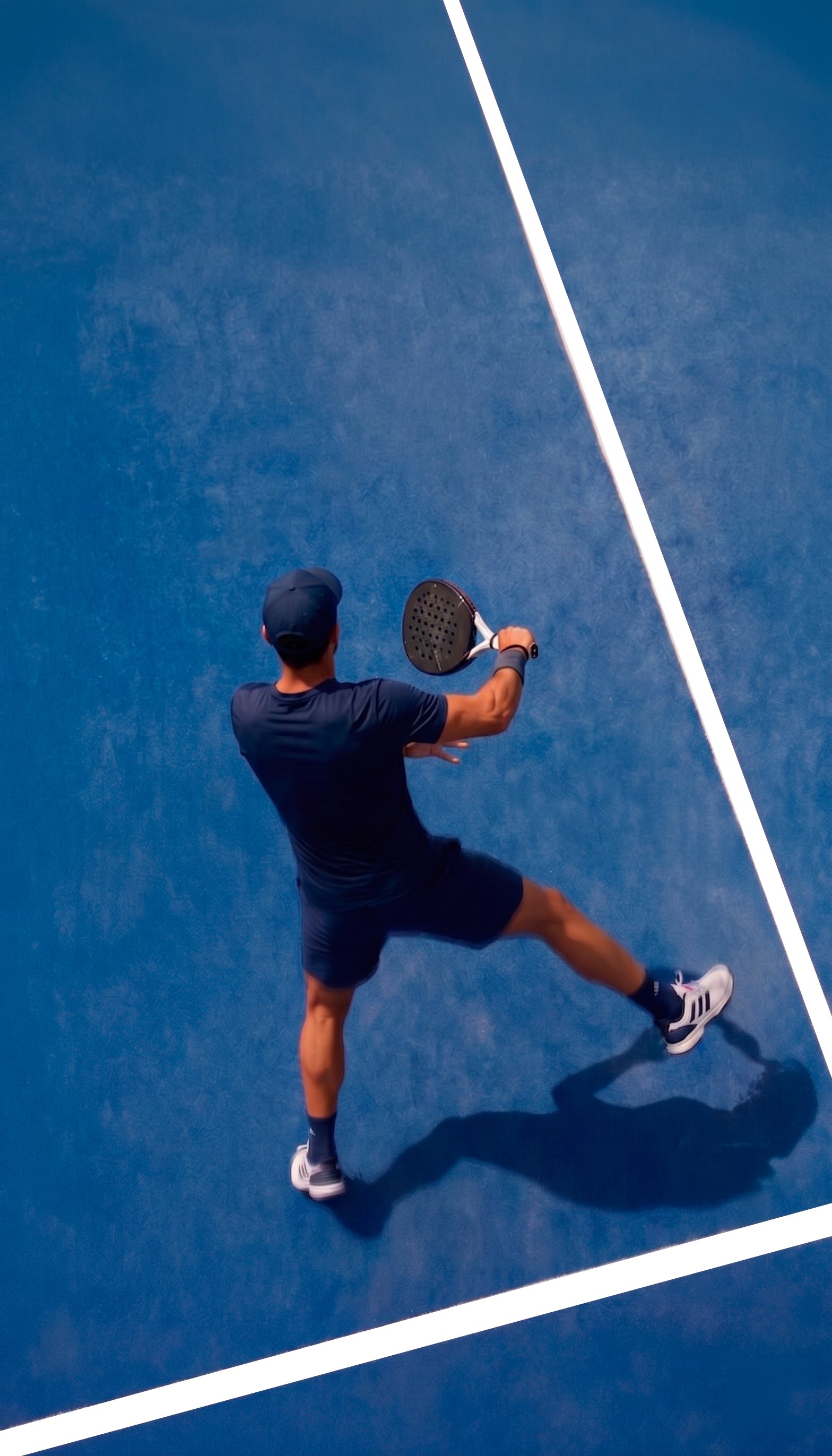 A padel player mid-rally on a deep blue padel court, seen from above.